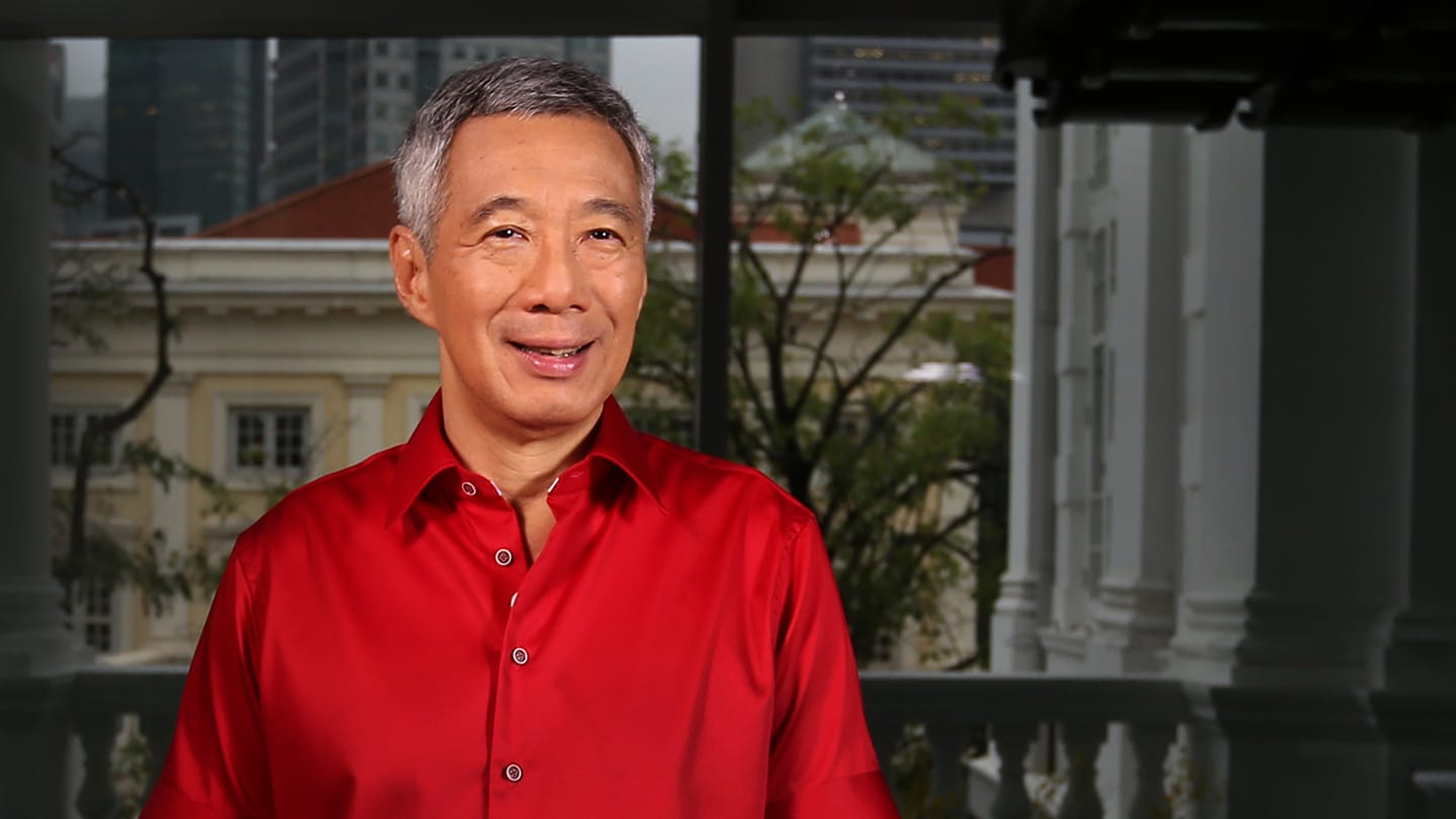 Lee Hsien Loong in red shirt, stands against a blurred cityscape backdrop.